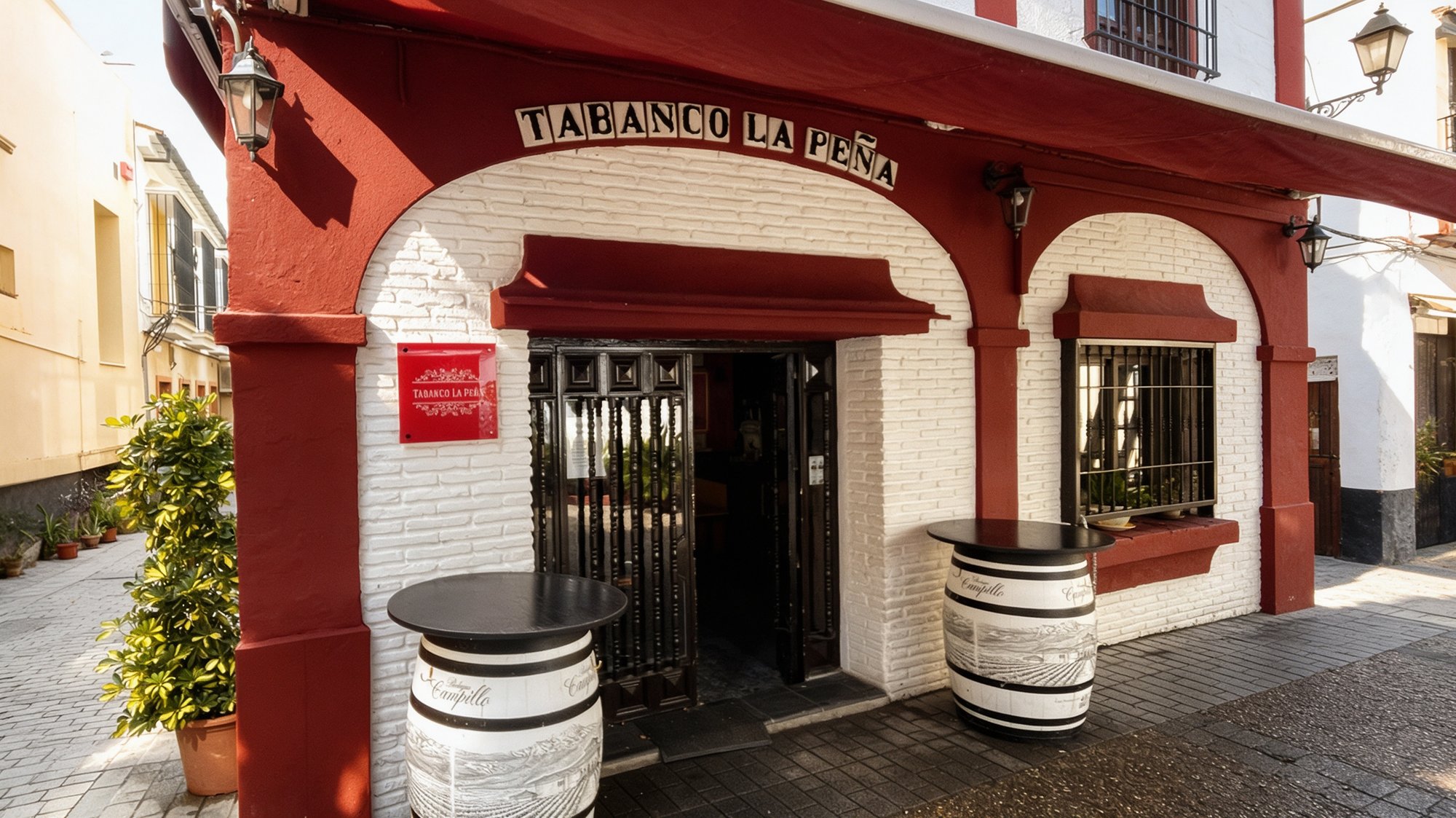 Fachada tradicional de tabanco andaluz con arcos rojos sobre muro encalado, rótulo de azulejos blancos que dice TABANCO LA PEÑA y barricas de jerez como mesas altas al sol de la tarde